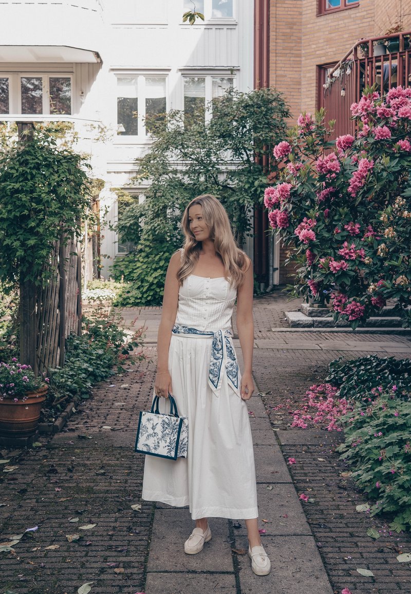 Femme en robe blanche et mocassins se tenant sur un chemin pavé, tenant un sac à main fleuri, entourée de buissons en fleurs et d'habitations.