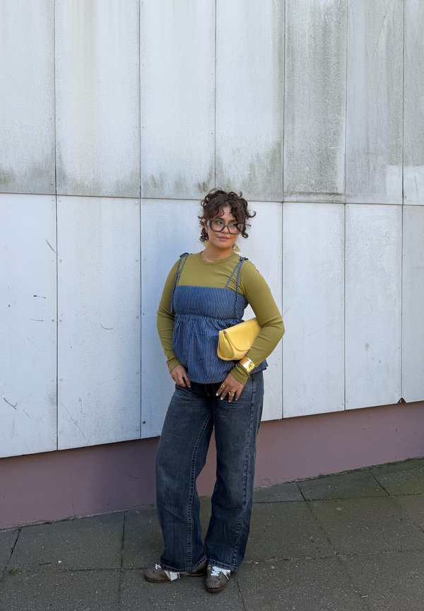 Persona con cabello rizado y gafas de pie contra una pared blanca, vistiendo una camiseta verde de manga larga, una camiseta sin mangas azul a rayas, jeans negros y sosteniendo un bolso de mano amarillo.