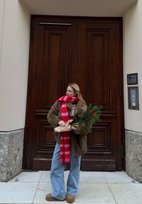 Woman wearing fur coat and red striped scarf, holding a wrapped evergreen branch, standing in front of large wooden double doors.