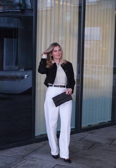 Chaqueta negra corta, suéter texturizado color crema, pantalones blancos de talle alto, bolso de mano negro y tacones negros puntiagudos frente a un fondo de vidrio.