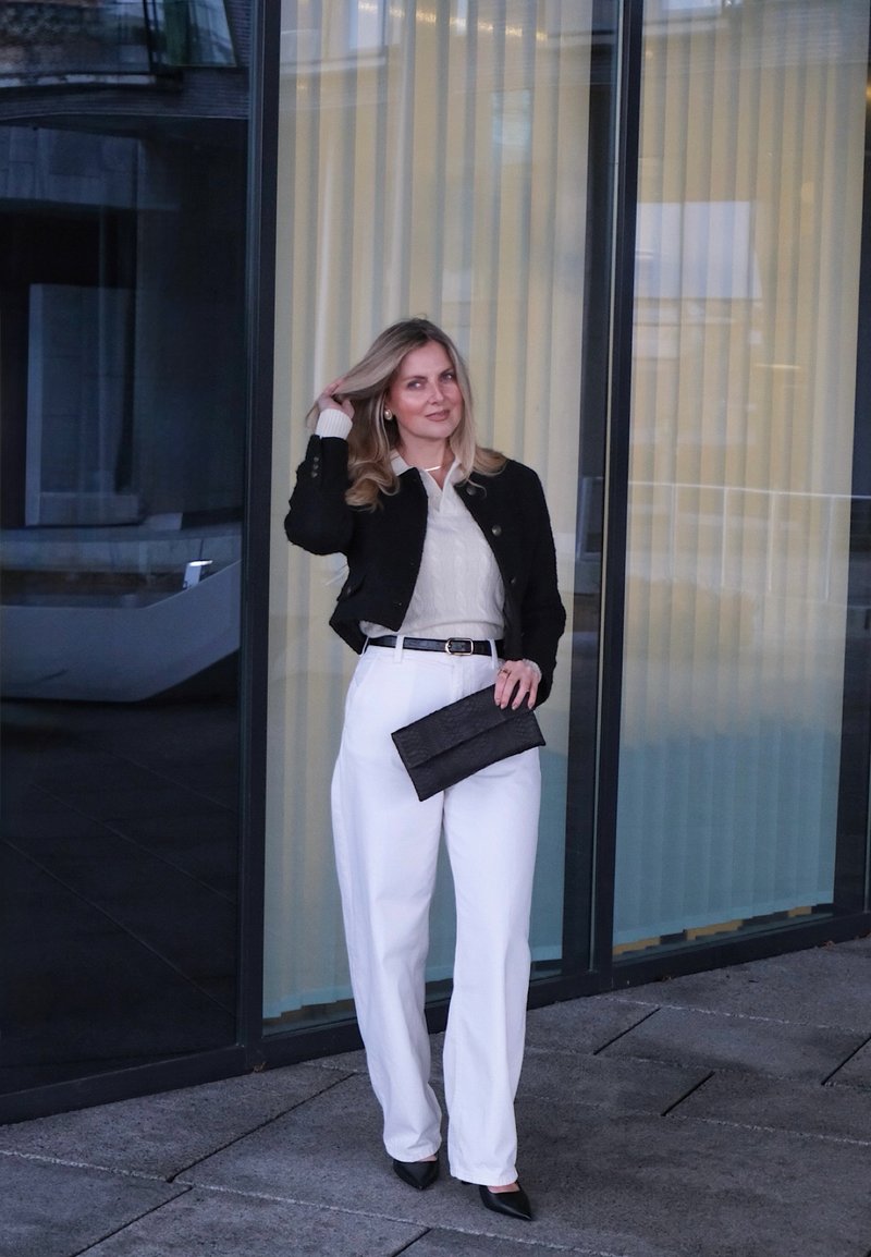 Giacca corta nera, maglione crema con texture, pantaloni bianchi a vita alta, pochette nera e décolleté nere a punta su sfondo di vetro.