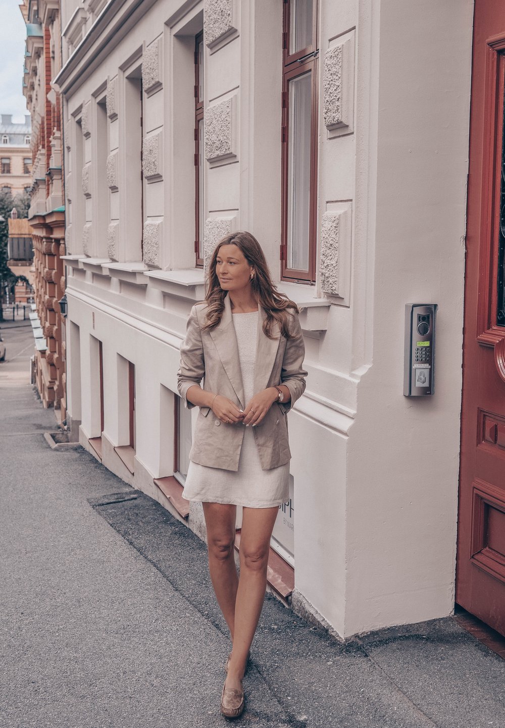 Beige blazer over een lichte jurk, staande op een stoep naast een textuurrijk wit gebouw en een rode deur, met een slanke intercom in de buurt.