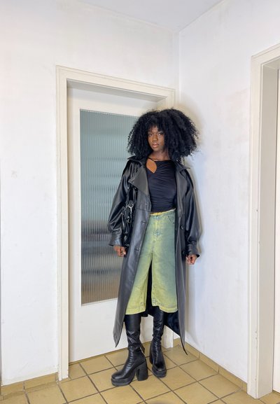 Abrigo de trench de imitación piel negra sobre una blusa negra, combinado con jeans acampanados de un verde claro y botas de plataforma negras hasta la rodilla. Incluye un bolso negro.