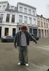 Άτομο που φοράει ένα καρό πουκάμισο πάνω από ένα λευκό T-shirt, φαρδιά ξεθωριασμένα τζιν και λε�υκά αθλητικά παπούτσια, στέκεται σε έναν δρόμο με κτίρια και σταθμευμένα αυτοκίνητα.