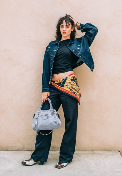 Chaqueta de mezclilla con acentos de estrellas, top negro, bufanda estampada en la cintura, pantalones oscuros, bolso gris y zapatos bicolores con punta afilada.