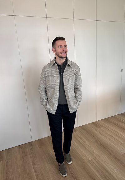 Hombre sonriente con las manos en los bolsillos, con chaqueta de cuadros, camisa oscura, pantalones negros y zapatos grises sin cordones, de pie en el interior sobre suelo de madera.