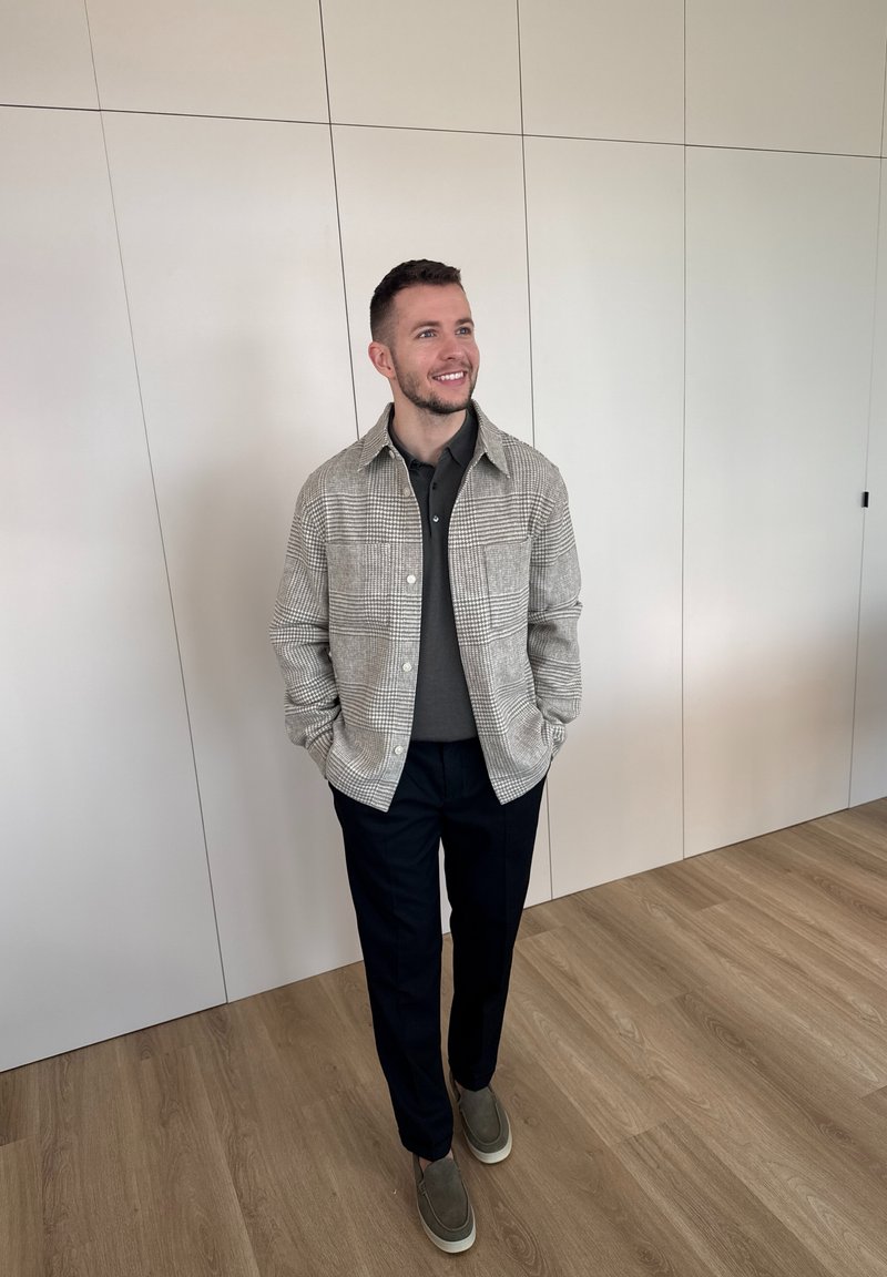 Άνδρας που χαμογελά με τα χέρια στις τσέπες, φοράει καρό σακάκι, σκούρο πουκάμισο, μαύρο παντελόνι και γκρι παπούτσια χωρίς κορδόνια, στέκεται σε εσωτερικό χώρο σε ξύλινο δάπεδο.