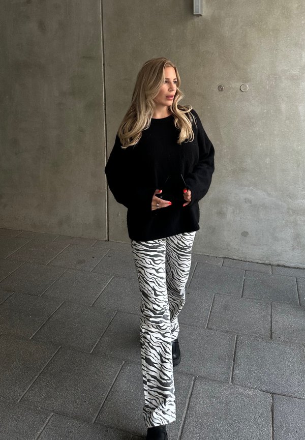 Black oversized sweater; white and gray zebra-patterned flared pants; black boots; standing on a stone surface with a concrete wall backdrop.