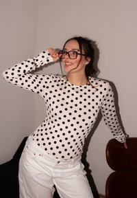 Γυναίκα που φοράει μαύρα γυαλιά και λευκό μακρυμάνικο πουκάμισο με μαύρα πουά, ποζάρει μέσα σε εσωτερικό χώρο, ακουμπώντας σε καφέ καρέκλα.