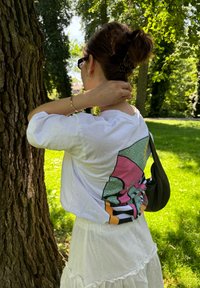 Weißes Shirt mit buntem Grafikdruck auf dem Rücken, getragen mit einem schichten weißen Rock. Haare zu einem Dutt frisiert, steht in der Nähe eines Baumes auf einer grasigen Fläche.