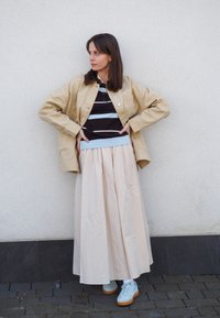 Giacca oversize beige, polo a righe nere e blu, gonna maxi plissettata crema e scarpe da ginnastica azzurre chiare con suola marrone. Sfondo semplice.