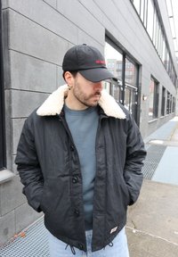 Homme portant une veste noire à col en peau lainée, un sweat-shirt bleu, un jean clair et une casquette noire, debout près d'un bâtiment moderne sur le trottoir.
