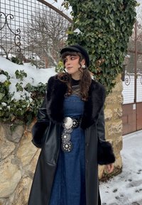Femme portant un manteau en cuir avec bordure en fourrure noire, robe en denim, large ceinture décorative et béret noir, debout à l'extérieur près de lierre enneigé et d'un mur en pierre.