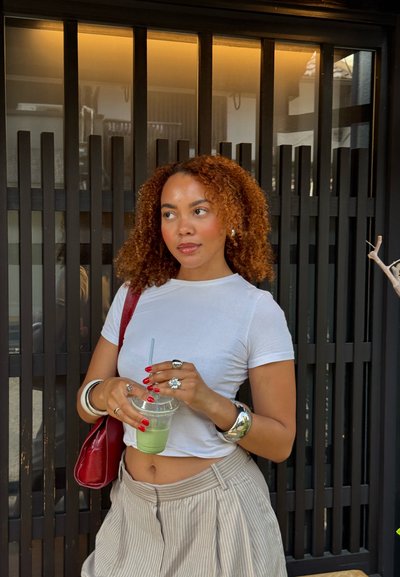 Top corto blanco de manga corta, pantalones de rayas de cintura alta, pulseras de plata, sosteniendo un vaso transparente con una bebida verde. El fondo presenta listones verticales negros.