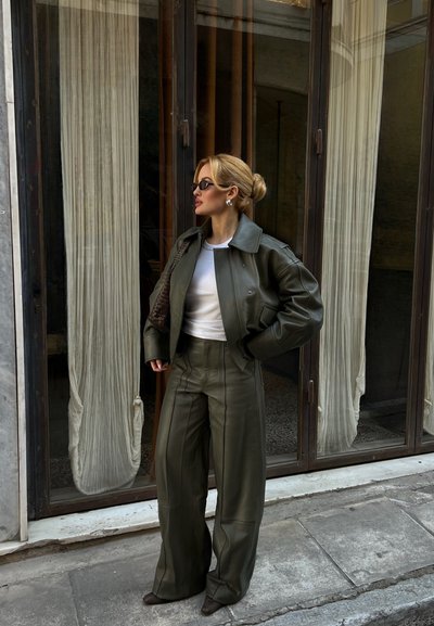 Mujer con moño rubio que lleva chaqueta y pantalones de cuero verde oscuro, camisa blanca, gafas de sol, de pie cerca de puertas de cristal con cortinas.