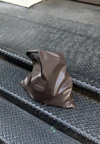 Bolsa tote castanha e brilhante, feita de material texturizado, amarrotada sobre degraus de metal preto e estriado. Apresenta uma alça única e costura mínima.
