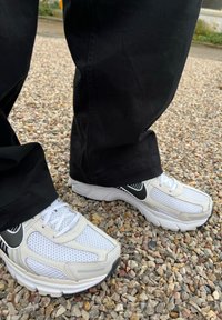 Λευκά αθλητικά παπούτσια με επάνω από διχτυωτό υλικό, μαύρες λεπτομέρειες και υφασμάτινες σόλες; φορεμένα με μαύρο παντελόνι σε μια επιφάνεια χαλικιού.