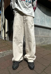 Jeans de corte largo, de um bege claro, com detalhes de costura visíveis, combinados com sapatilhas pretas. O fundo inclui um pavimento e a fachada de um edifício.