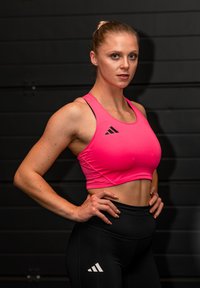 Atleetlik naine blondide juustega, kandes roosat spordirinnahoidjat ja musti retuuse, seisab käed puusas tume seina taustal.