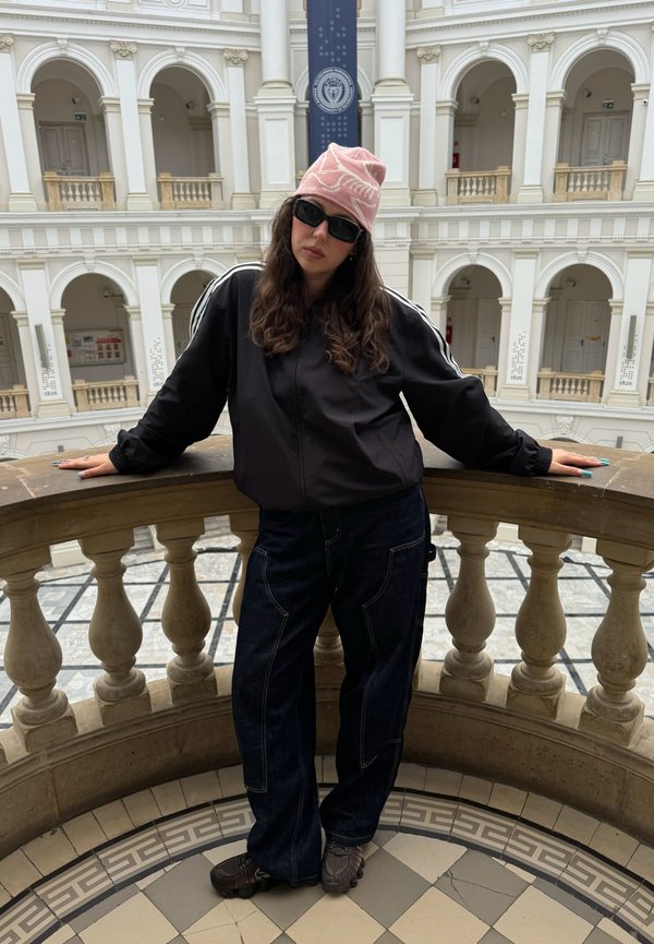 Mladá žena s okuliarmi na slnko a ružovým klobúkom sa opiera o kamenné zábradlie vo veľkej budove s vysokými oblúkmi a balkónmi.