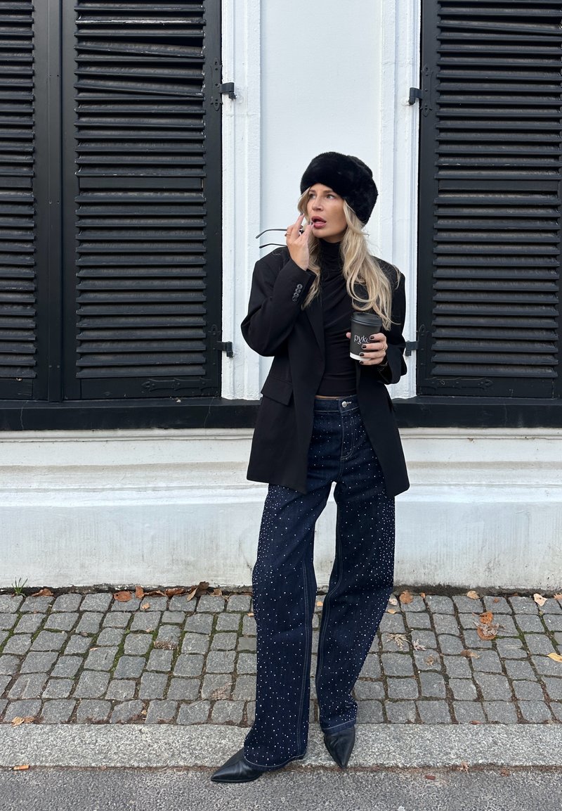 Veste blazer noire oversize sur un col roulé noir, jean bleu foncé avec des ornements blancs, et un chapeau en fourrure noir. Tenant une tasse de café.