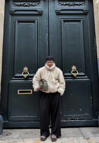 Manteau en fausse fourrure crème, surdimensionné avec un col montant, assorti à un pantalon noir à jambes larges. Tenant un sac vert olive, debout devant des portes en bois sombre.