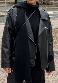 Personne portant une veste en cuir noire, une écharpe noire et un pull noir, debout dehors dans une rue pavée de pierre.