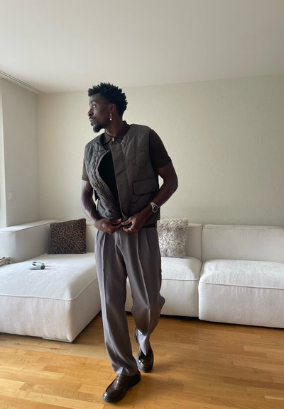 Hombre abotonándose un chaleco acolchado oscuro sobre una camisa marrón, vistiendo pantalones grises y zapatos marrones, de pie en la sala de estar junto a un sofá blanco.