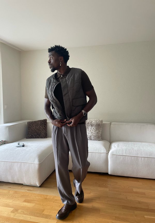 Man die een donker doorgestikt vest dichtknopt over een bruin shirt, draagt een grijze broek en bruine schoenen, staand in de woonkamer met een witte bank.