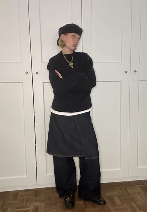 Suéter negro de punto, camisa blanca con capas, falda de mezclilla con corte asimétrico, pantalones oscuros de pierna ancha, gorra negra, collares dorados con capas.
