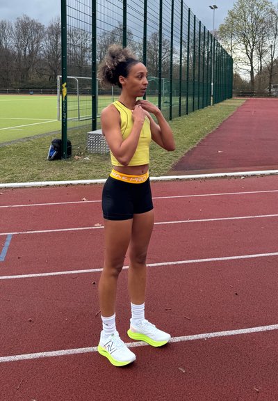 Mujer atlética con top corto amarillo y pantalones cortos negros ajustándose un collar mientras está de pie en la pista roja de atletismo cerca de la valla del campo deportivo.