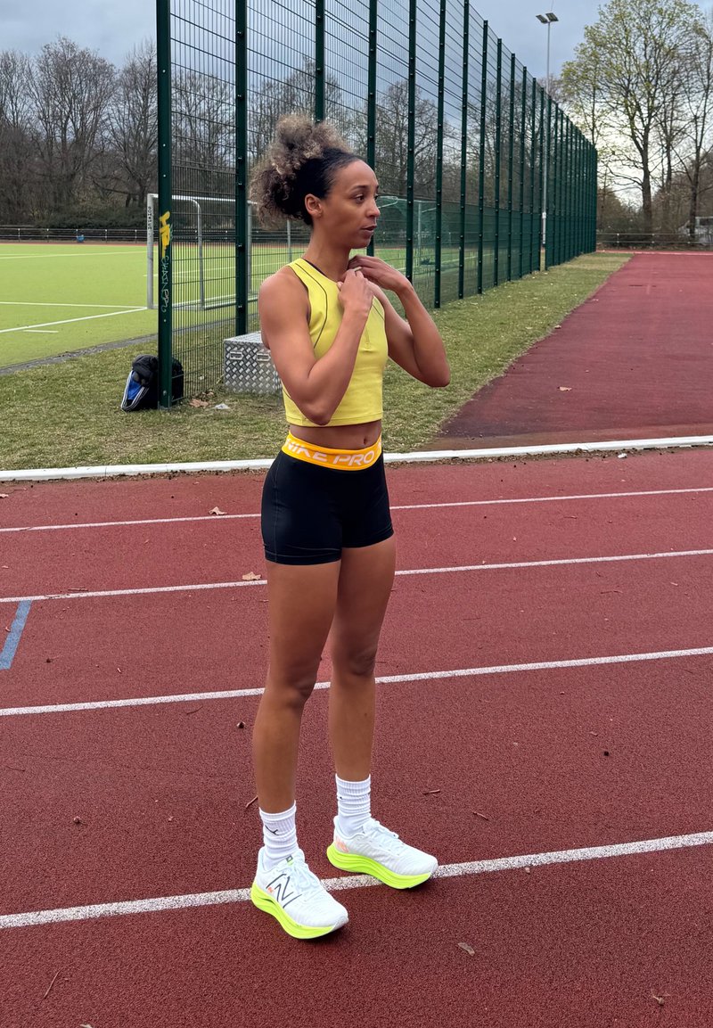 Femme athlétique en crop top jaune et short noir ajustant un collier tout en se tenant sur la piste rouge près de la clôture du terrain de sport.