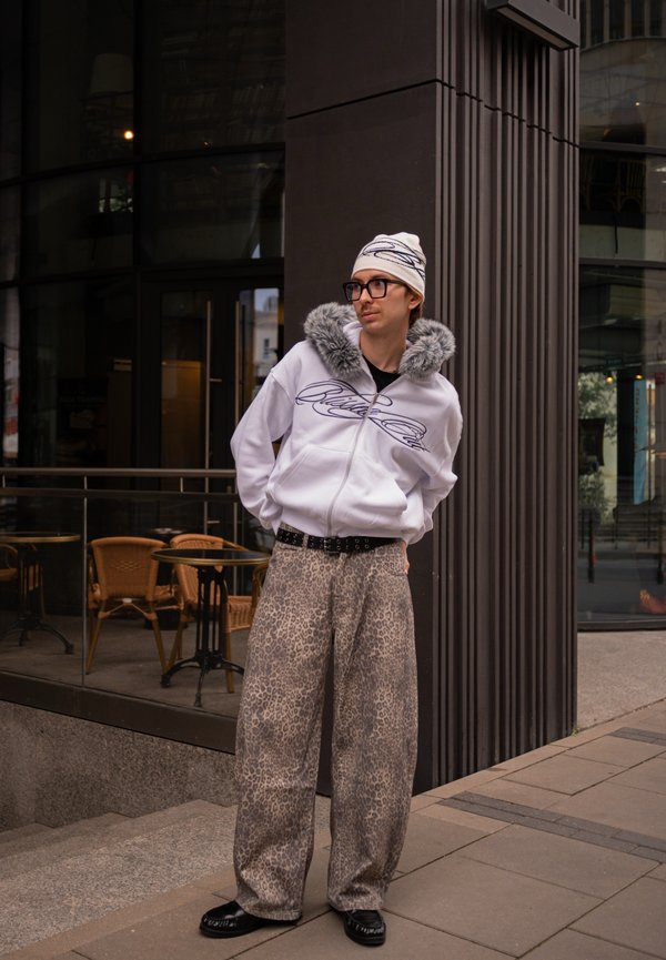Una persona con gafas, una chaqueta con ribete de pelo blanco, pantalones estampados y un gorro de punto está de pie frente a una cafetería.