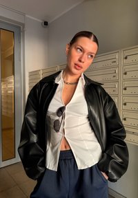 Νεαρή γυναίκα με μαλλιά χτενισμένα προς τα πίσω, ντυμένη με μαύρο δερμάτινο μπουφάν πάνω από ένα λευκό πουκάμισο με μερικά κουμπιά ανοιχτά και μπλε παντελόνι, στέκεται δίπλα σε γραμματοκιβώτια.