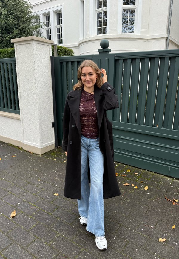 Un manteau noir croisé porté sur un top en dentelle bordeaux et un jean évasé bleu clair, associé à des baskets blanches, posant en extérieur près d'une porte verte.