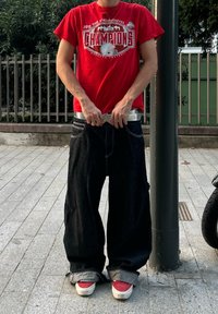 Laza, sötét farmernadrág, amelyen látható márkajelzések vannak, ezüst övvel és piros pólóval, amely sportgrafikát ábrázol. Cserélt felületen áll.