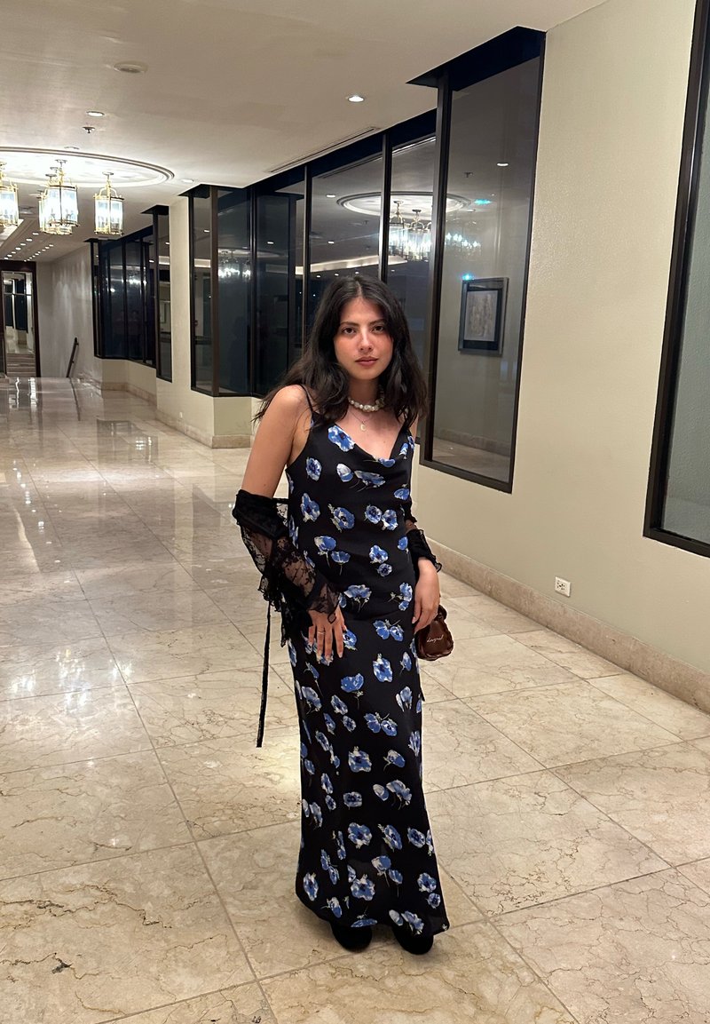 Žena v černých květovaných šatech s průhledným černým šálem stojí na chodbě s mramorovou podlahou, skleněnými okny a stropními světly.