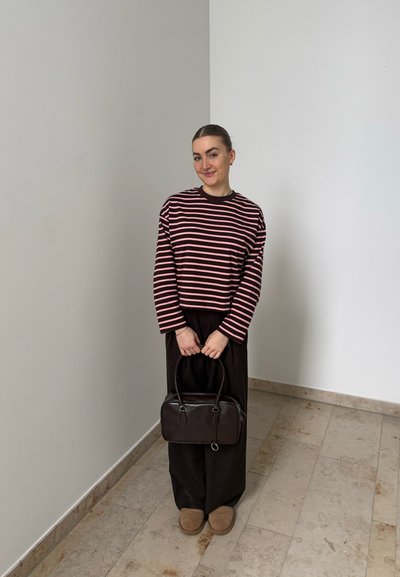 Mujer con una camiseta de manga larga a rayas y pantalones negros, sosteniendo un bolso oscuro, de pie sobre un suelo de baldosas frente a una pared lisa.