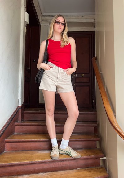 Top sin mangas rojo, pantalones cortos de mezclilla beige, calcetines tobilleros blancos, zapatillas verdes y marrones, sosteniendo un bolso negro texturizado, fondo de escalera de madera.