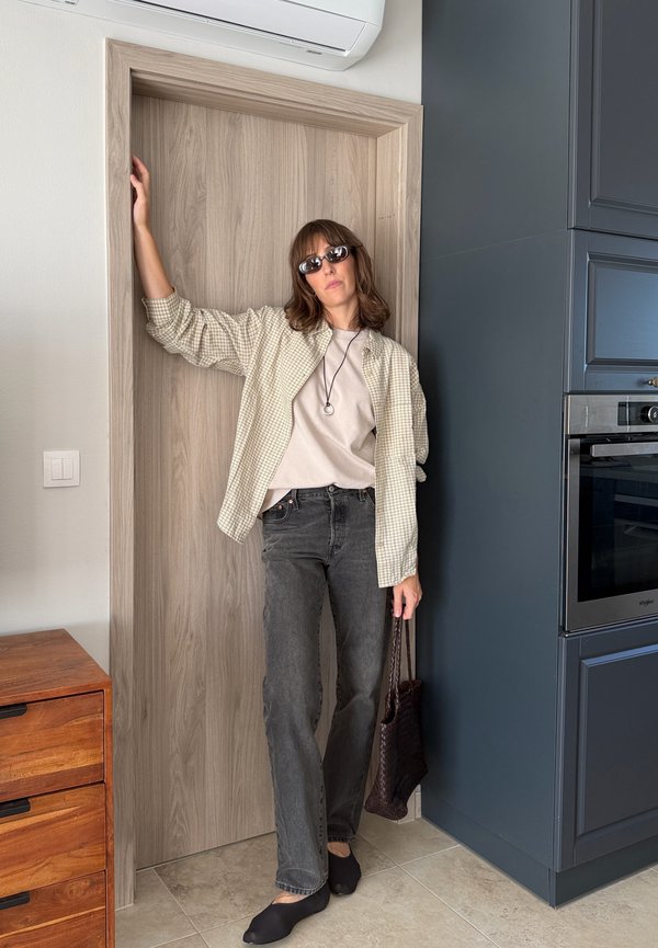 Mujer con gafas de sol, chaqueta a cuadros clara, camisa blanca y vaqueros negros, está en el marco de una puerta de la cocina sosteniendo un bolso oscuro tejido.