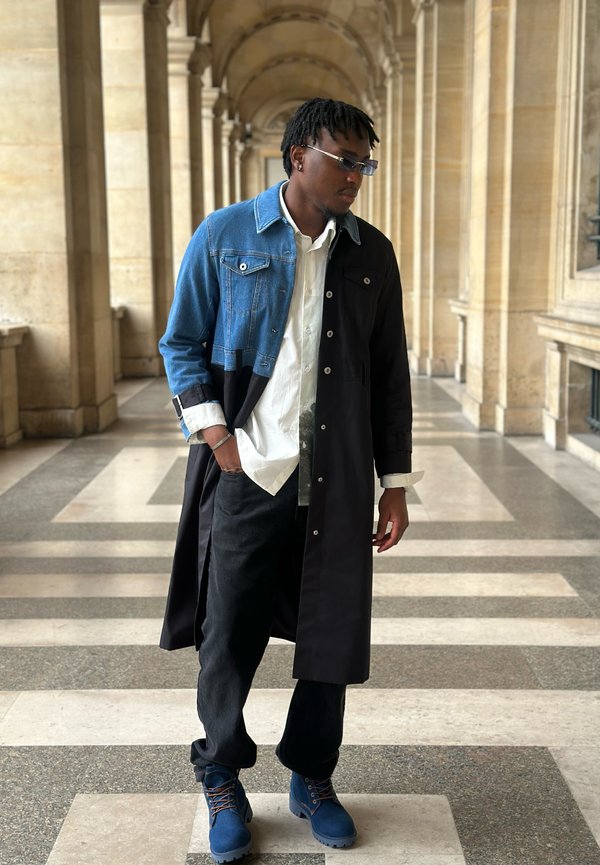 Homme avec des dreadlocks courtes portant des lunettes de soleil, un manteau moitié en denim moitié noir, une chemise blanche, un pantalon noir et des bottes bleues marchant dans un couloir en pierre avec des arches.