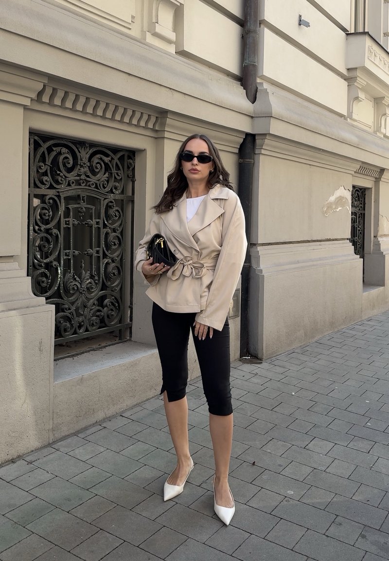 Femme portant une veste beige avec ceinture, pantalon noir court, talons blancs et lunettes de soleil, debout sur un trottoir carrelé près d'une rambarde ornée et d'un bâtiment beige.