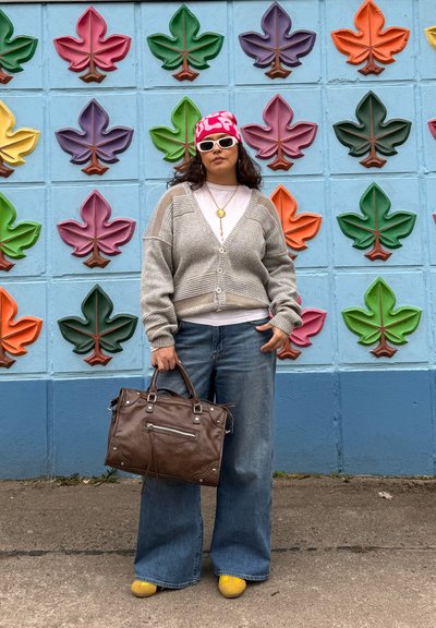 Mujer con un pañuelo rosa estampado en la cabeza, gafas de sol blancas, cárdigan gris, vaqueros anchos, zapatos amarillos, sosteniendo un bolso marrón, de pie junto a una pared de hojas coloridas.