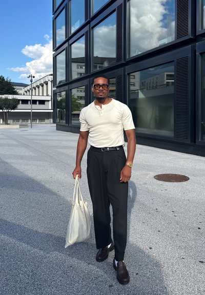 Camisa henley de manga corta blanca, pantalones de cintura alta negros, mocasines marrones, sosteniendo una bolsa tote beige, de pie sobre un pavimento texturizado.