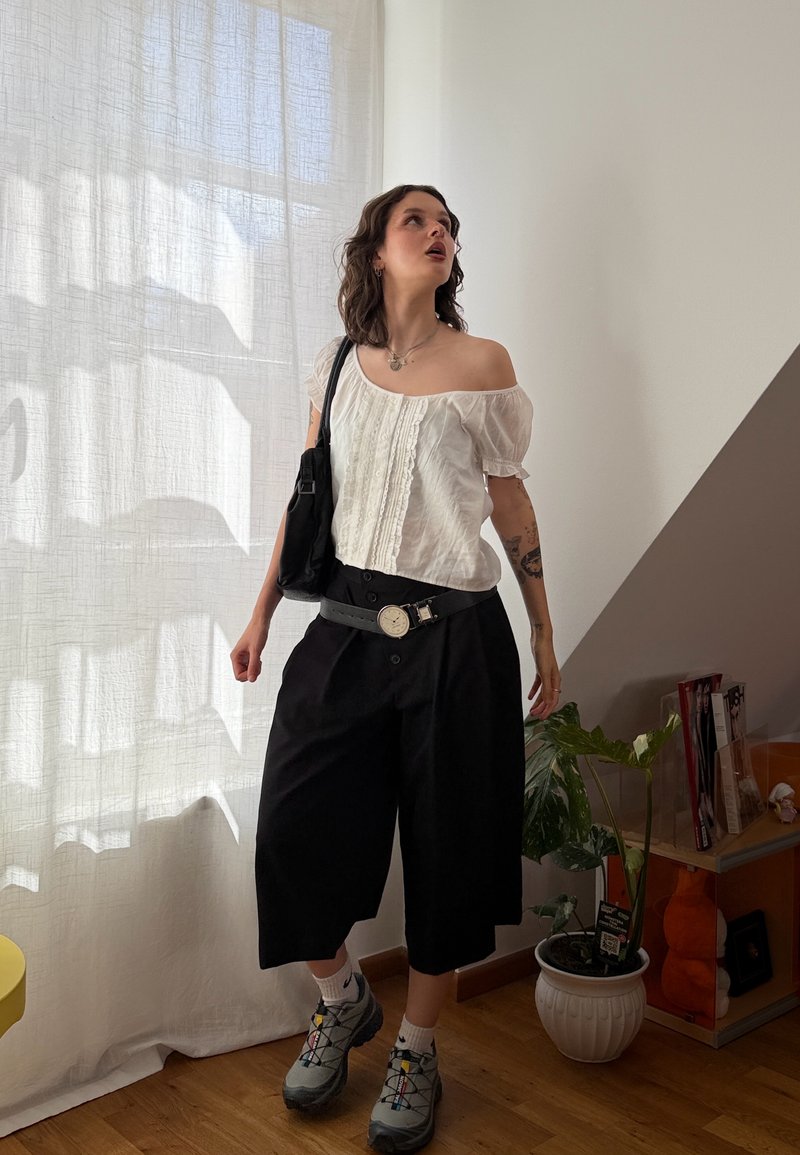 Woman wearing white off-shoulder top, black wide-leg pants, gray sneakers, and a black shoulder bag standing indoors near a window with sheer curtains.