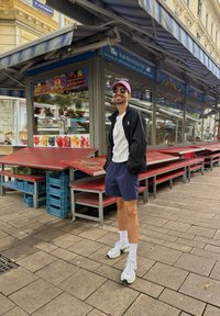 Svart jacka, vit skjorta, marinblå shorts, rosa keps, solglasögon och vita sneakers. Röda bord och blå lådor i en marknadsmiljö.