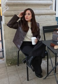 Kvinne med langt brunt hår iført brun skinnjakke, svarte bukser og støvler sitter på en stol og holder en kaffekopp ved en steinvegg og bord.
