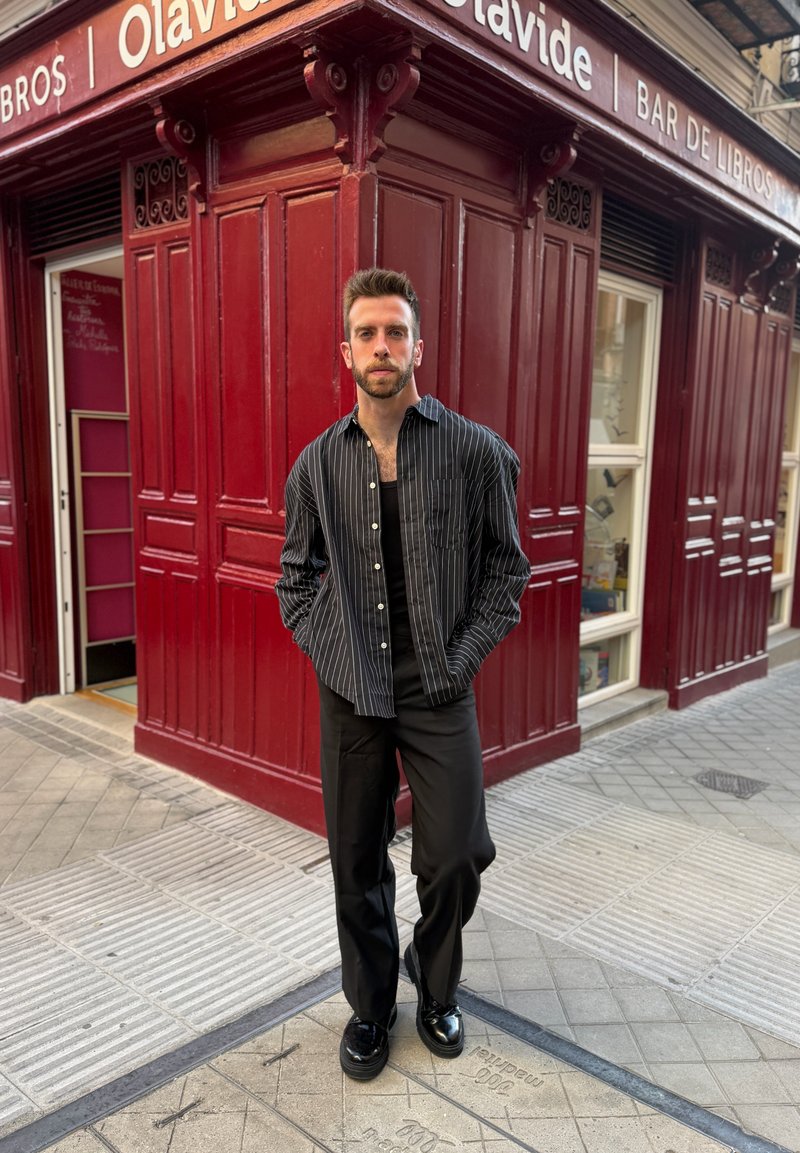 Homme en chemise et pantalon noirs à fines rayures debout sur le trottoir devant un bâtiment rouge avec le panneau "Olavide Bar de Libros."