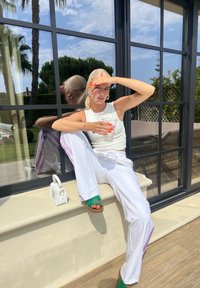 Λευκή κοντή μπλούζα, χαλαρές λευκές παντελόνες με μωβ ρίγες, πράσινες σαγιονάρες, κρατώντας ένα διάφανο ποτήρι, καθισμένη δίπλα σε γυάλινο παράθυρο με αντανακλάσεις.