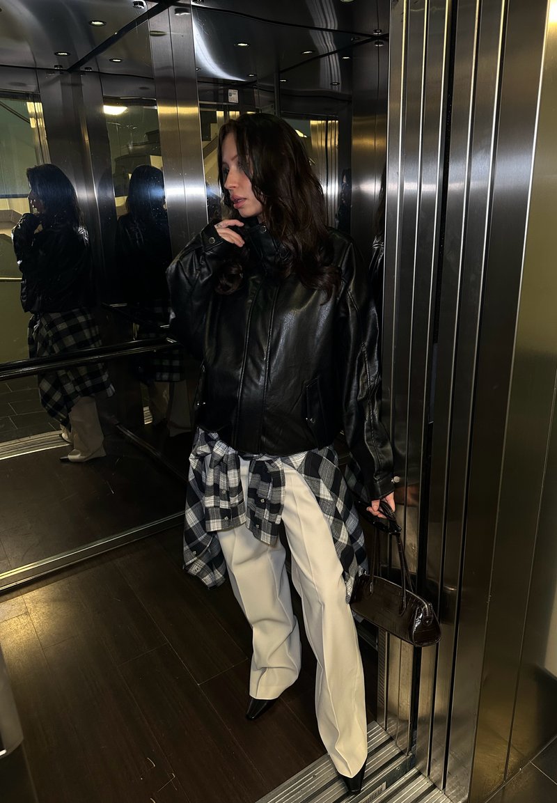 Black leather jacket, white wide-leg trousers, plaid shirt tied around the waist, and a small dark handbag. Reflections in the lift mirrors.